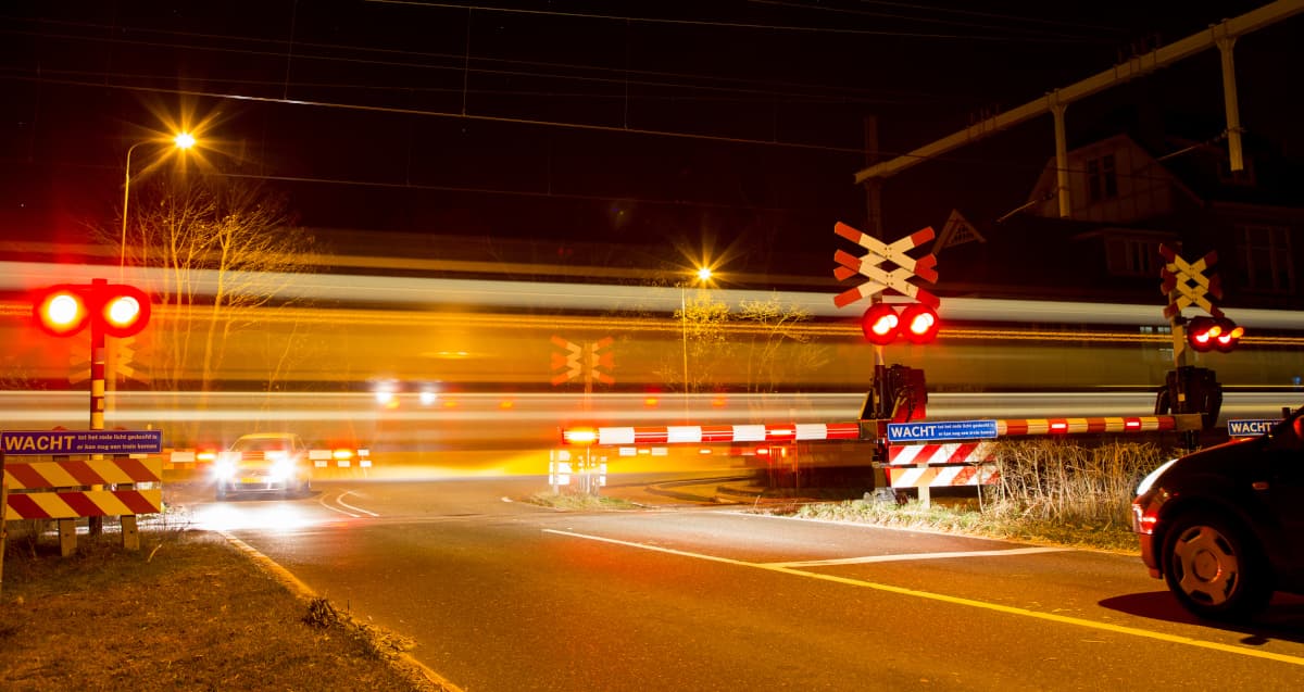 Trackside level crossing boom barrier in the lowered position at dusk, red-and-white retroreflective boom arm across the road with LED lamps lit, the barrier machine drive housing and counterweight at the post base and the signalling location case visible behind.