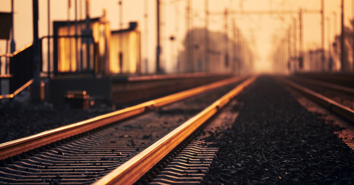 A trackside signalling location case at dusk beside the running rails, a fibre and antenna cable rising from it towards a distant lit building on the horizon, suggesting a data path from the wayside to a central platform under a deep blue twilight sky.